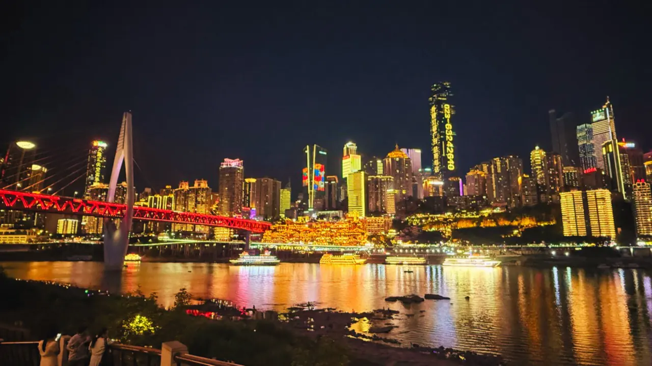 Chongqing at night — the lit Hongyadong complex on the riverbank below a wall of neon towers.
